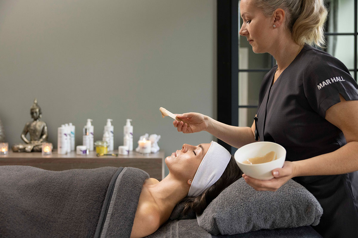 spa therapist applies face mask to a woman lying down on a massage table wrapped in a body towel and a large white headband