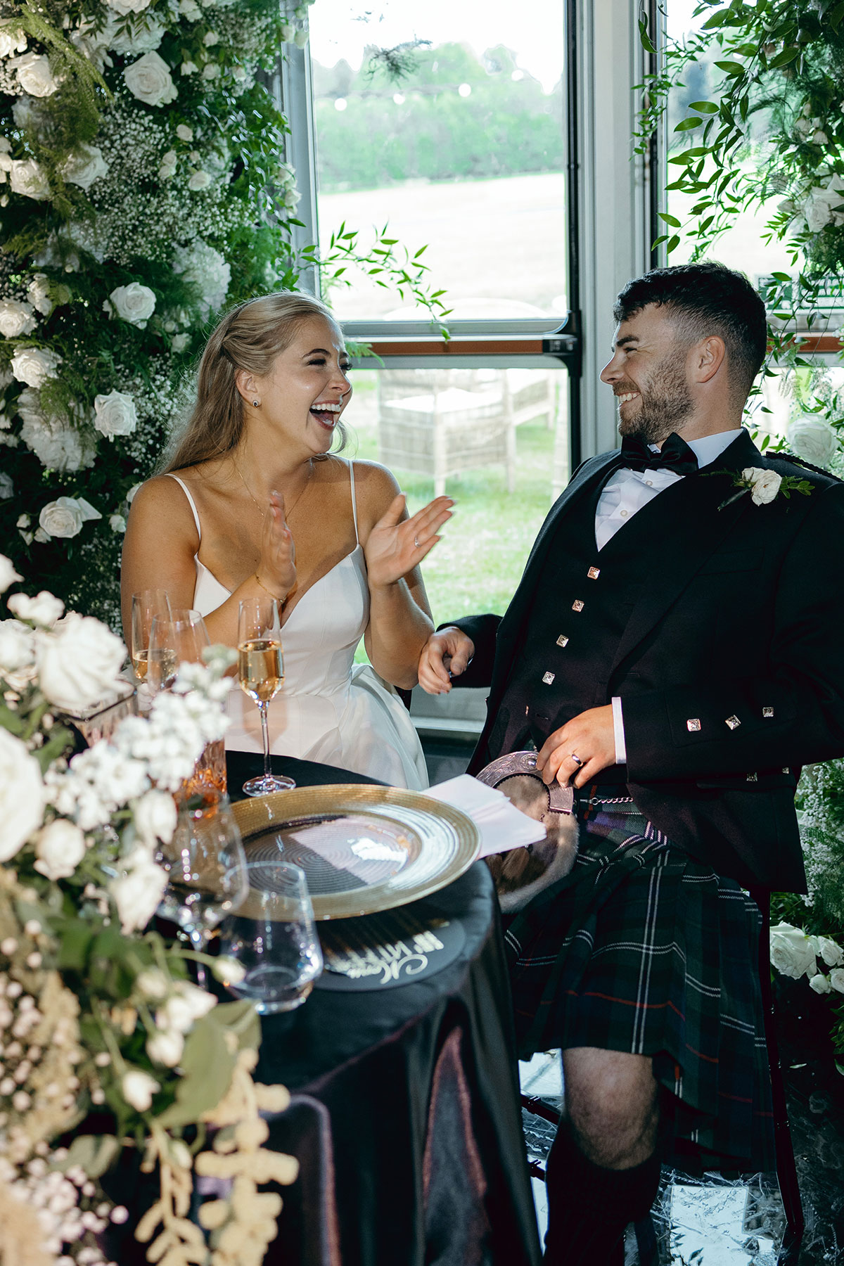 Bride and groom laugh together during wedding speeches surrounded by white florals at Dundas Castle.