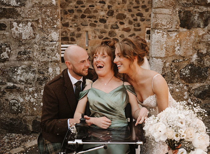 a bride and groom pose against a stone wall with a smiling person wearing a green dress who is sitting in a wheelchair. The bride is holding a large bouquet of white flowers