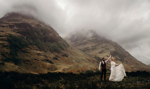 A bride and groom dancing together on a hilltop with other hills covered in mist in the background