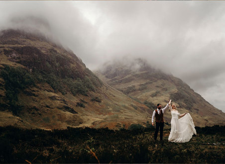 A bride and groom dancing together on a hilltop with other hills covered in mist in the background