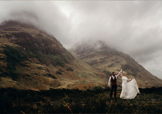 A bride and groom dancing together on a hilltop with other hills covered in mist in the background