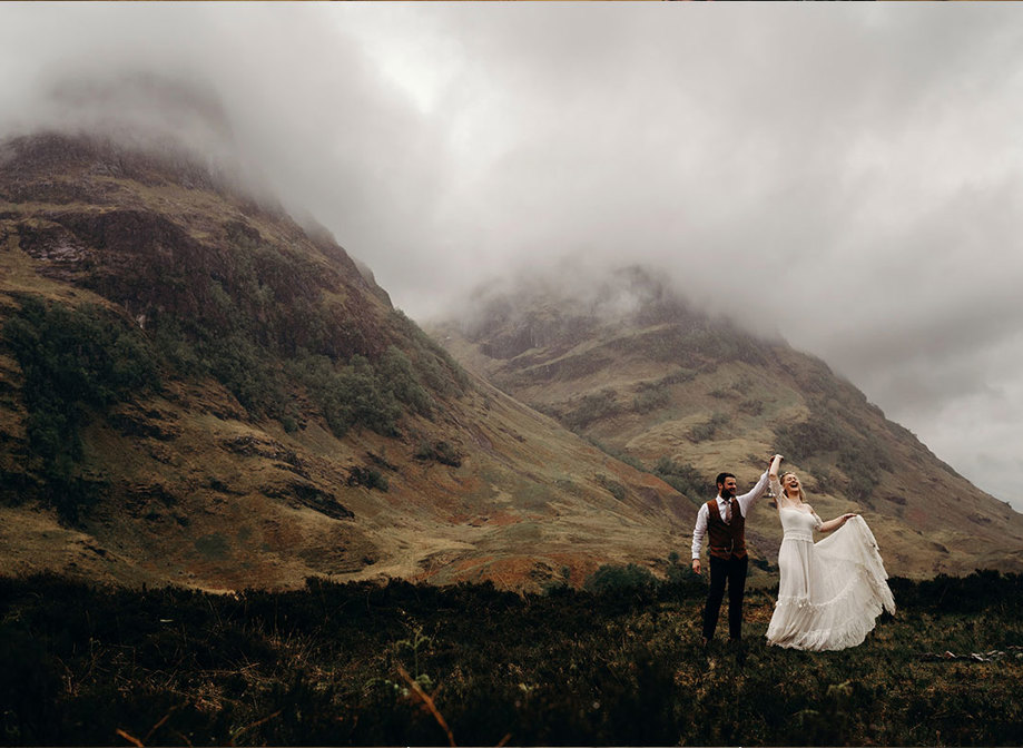 A bride and groom dancing together on a hilltop with other hills covered in mist in the background