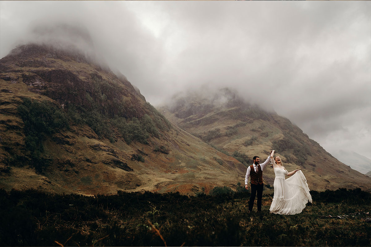 A bride and groom dancing together on a hilltop with other hills covered in mist in the background