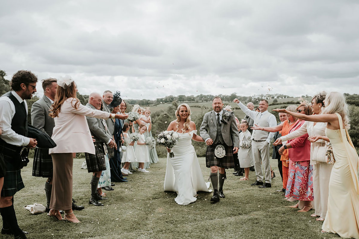 Bride and groom walk through an outdoor confetti aisle surrounded by guests at their Scottish wedding.