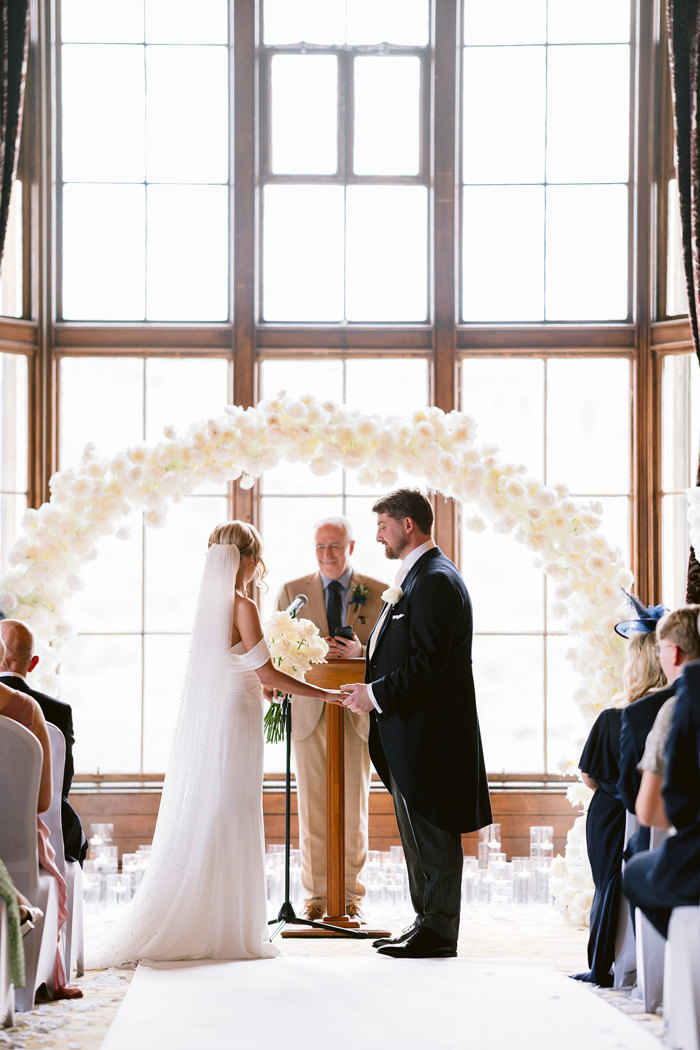 A bride and groom holding hands under a flower arch during a wedding ceremony at Mar Hall.