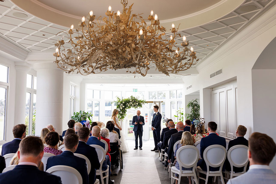 A wedding ceremony at the Old Course Hotel, in an elegant room with a large, artistic chandelier, as guests seated in rows look toward the couple standing at the altar