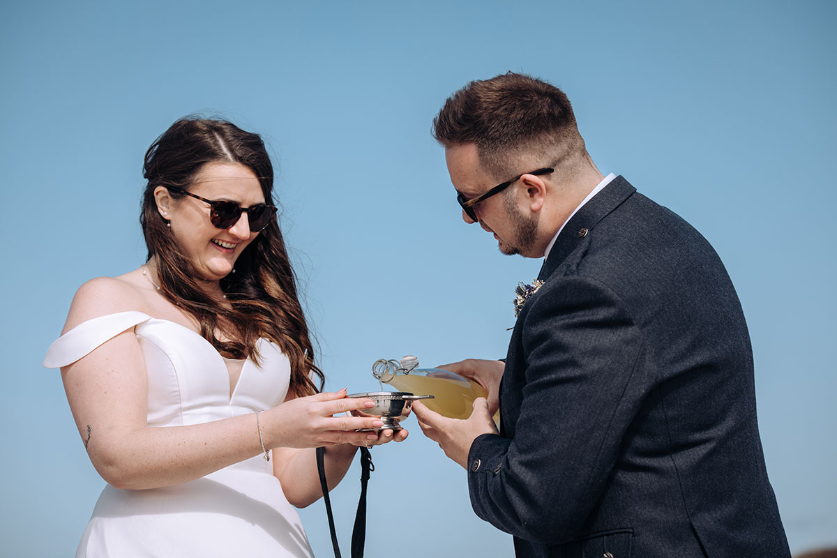 Bride and groom pour artisan limoncello into a silver quaich during their handfasting ceremony on the beach.