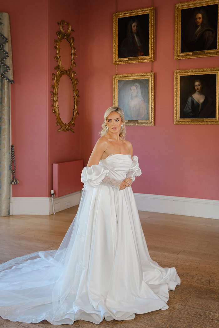 A bride wearing a strapless ballgown dress with added puffy sleeves, while standing in a large room with paintings on the wall behind her