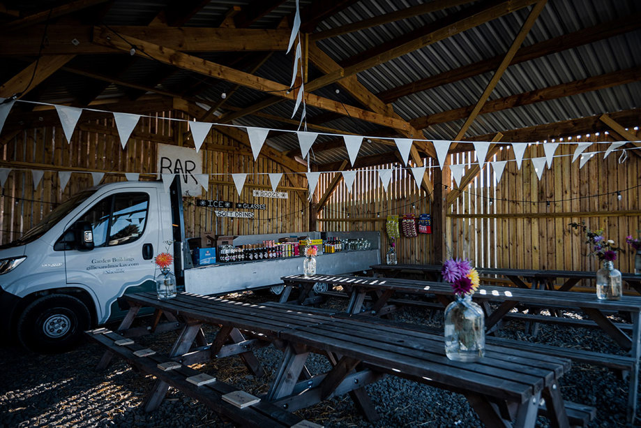 A Wooden Building Hung With White Bunting And White Flatbed Van And Long Picnic Tables And Benches Inside