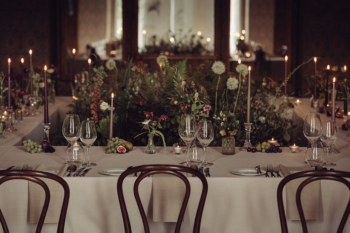 Candlelit wedding dining table set with white linen, layered glassware and abundant foliage centrepieces in a historic ballroom