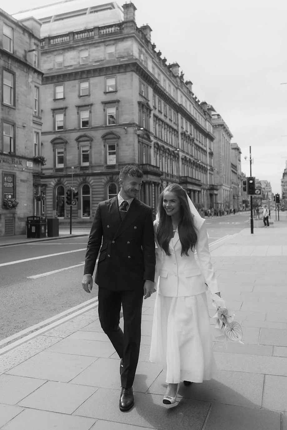 black and white photo of a couple holding hands walking down city street, man in dark suit and woman in white jacket over a dress, a veil in her hair and lilies in her hand