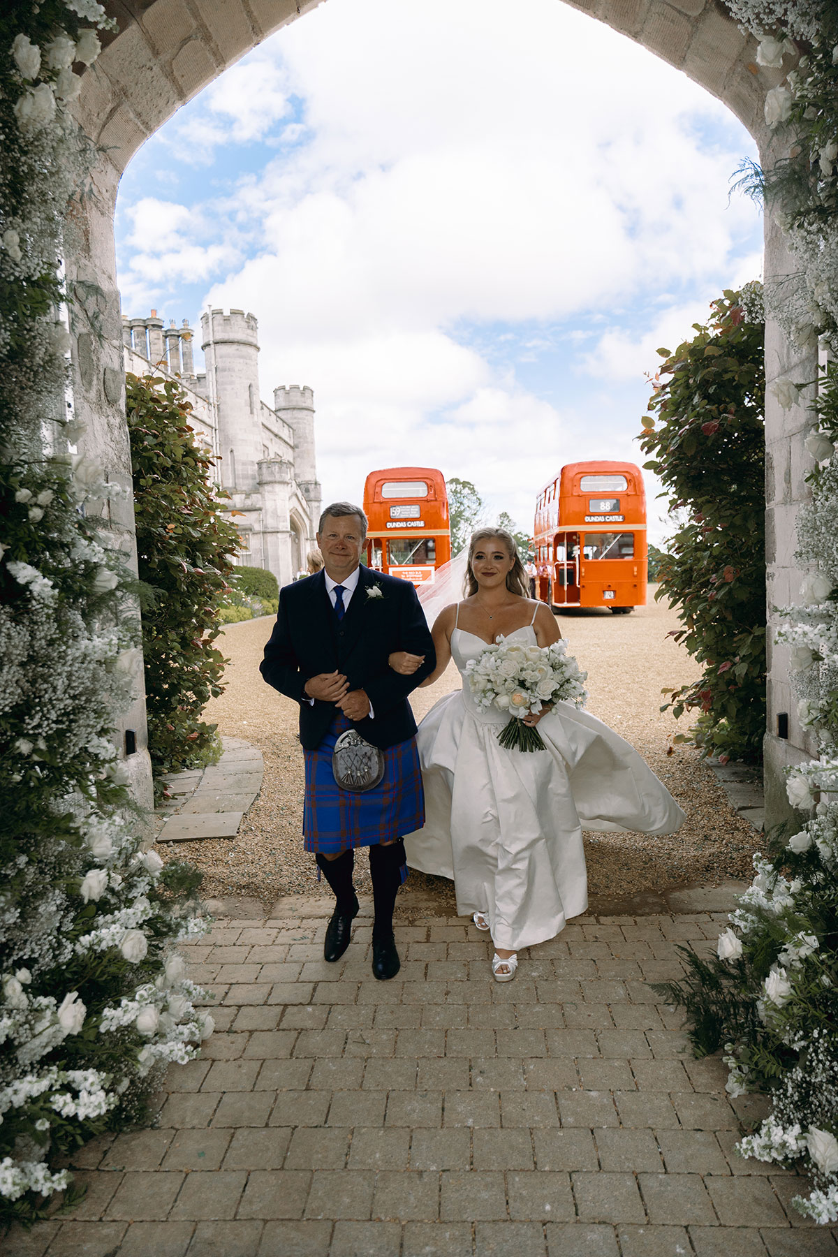 Bride walks through Dundas Castle archway with her father, red London buses in background.