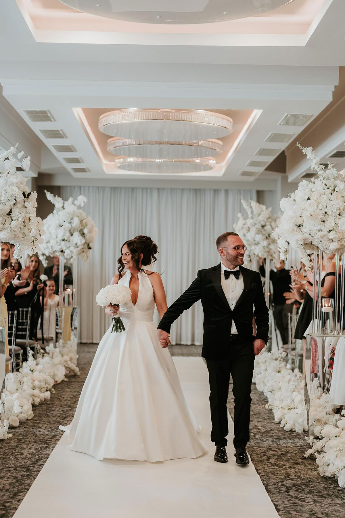 Bride and groom walking up the aisle after their ceremony at Riverside Lodge Hotel, with white floral arrangements lining the walkway and guests applauding.