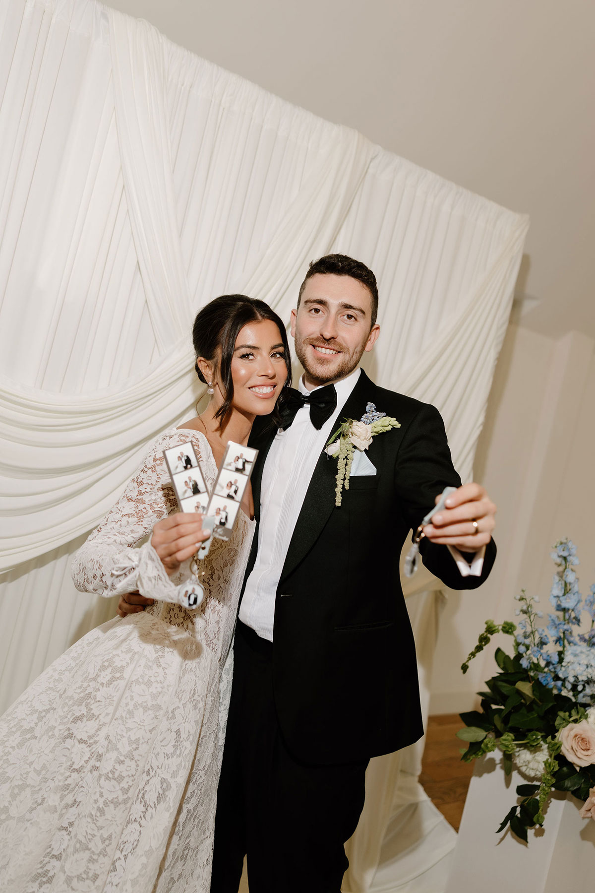 Bride and groom holding photo booth strips in front of a draped backdrop