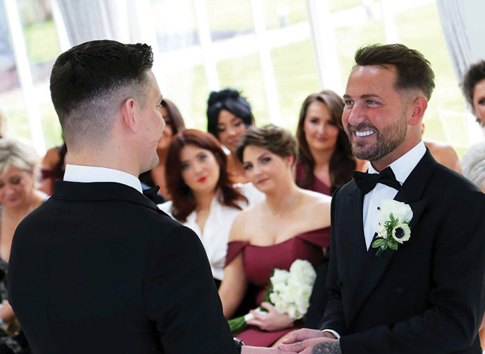 a groom wearing a smart black suit and bow tie smiles and holds hands with another groom during their wedding ceremony as seated wedding guests in the background look on