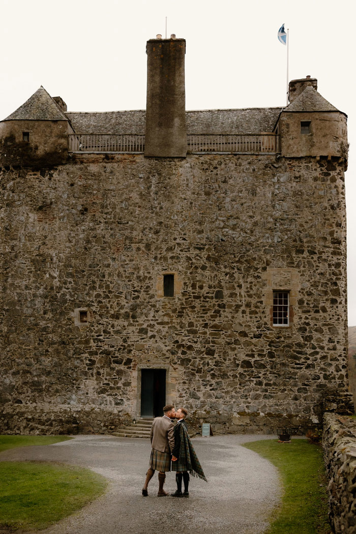 two grooms wearing kilts kissing outside Neidpath Castle