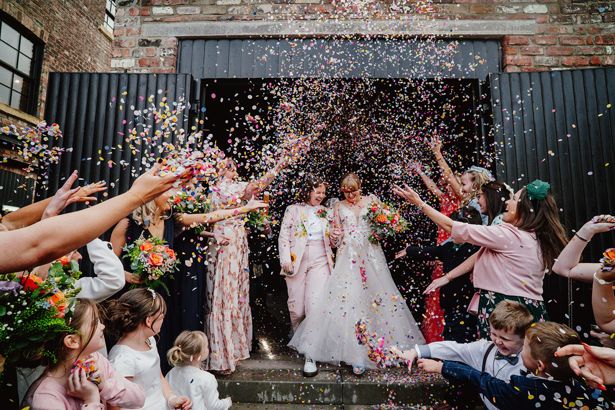 Newlywed brides walking out of the Engine Works as guests shower them with colourful confetti during their vibrant wedding exit.
