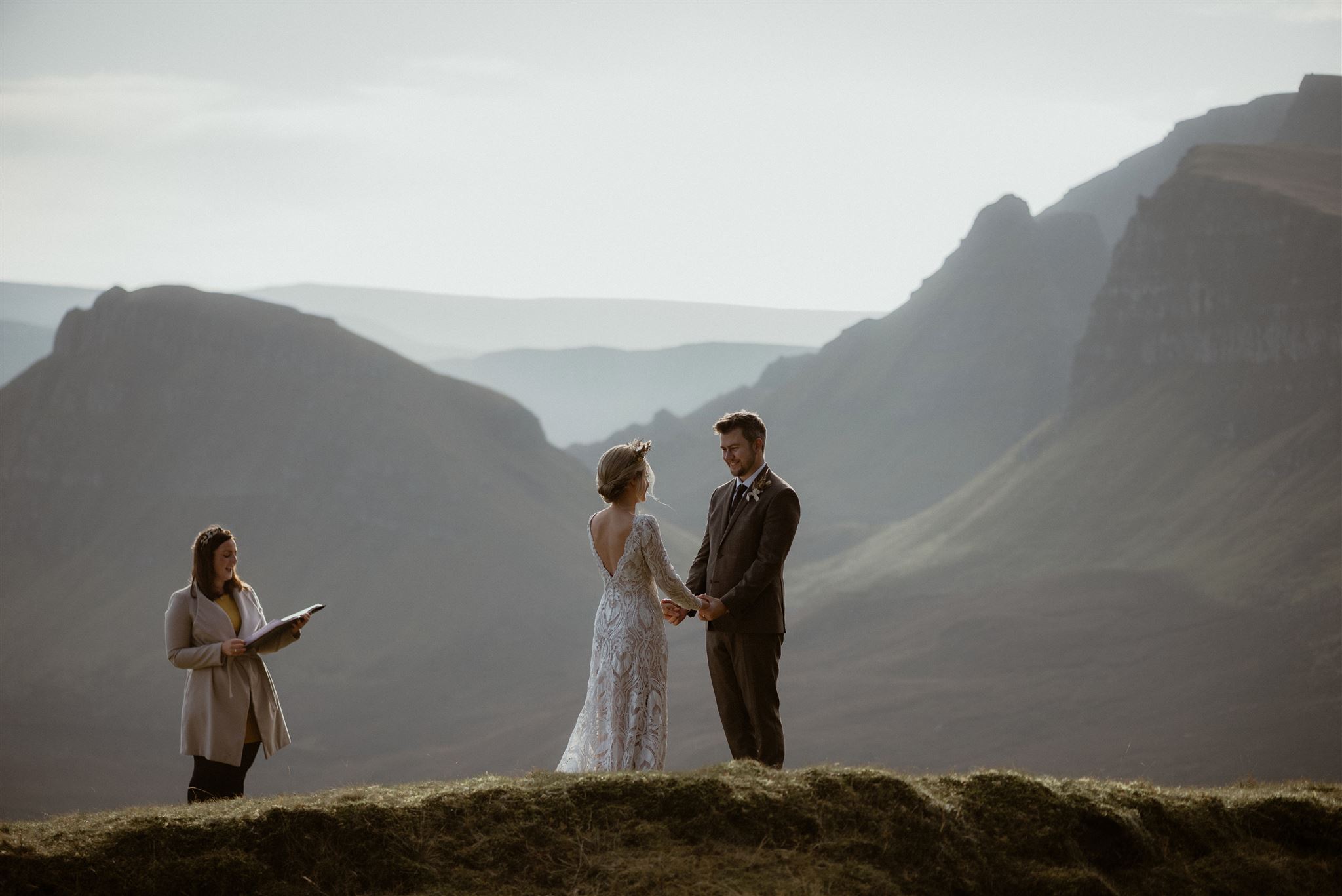 bride and groom hold hands on top of scottish mountain as humanist ceremony reads from book next to them