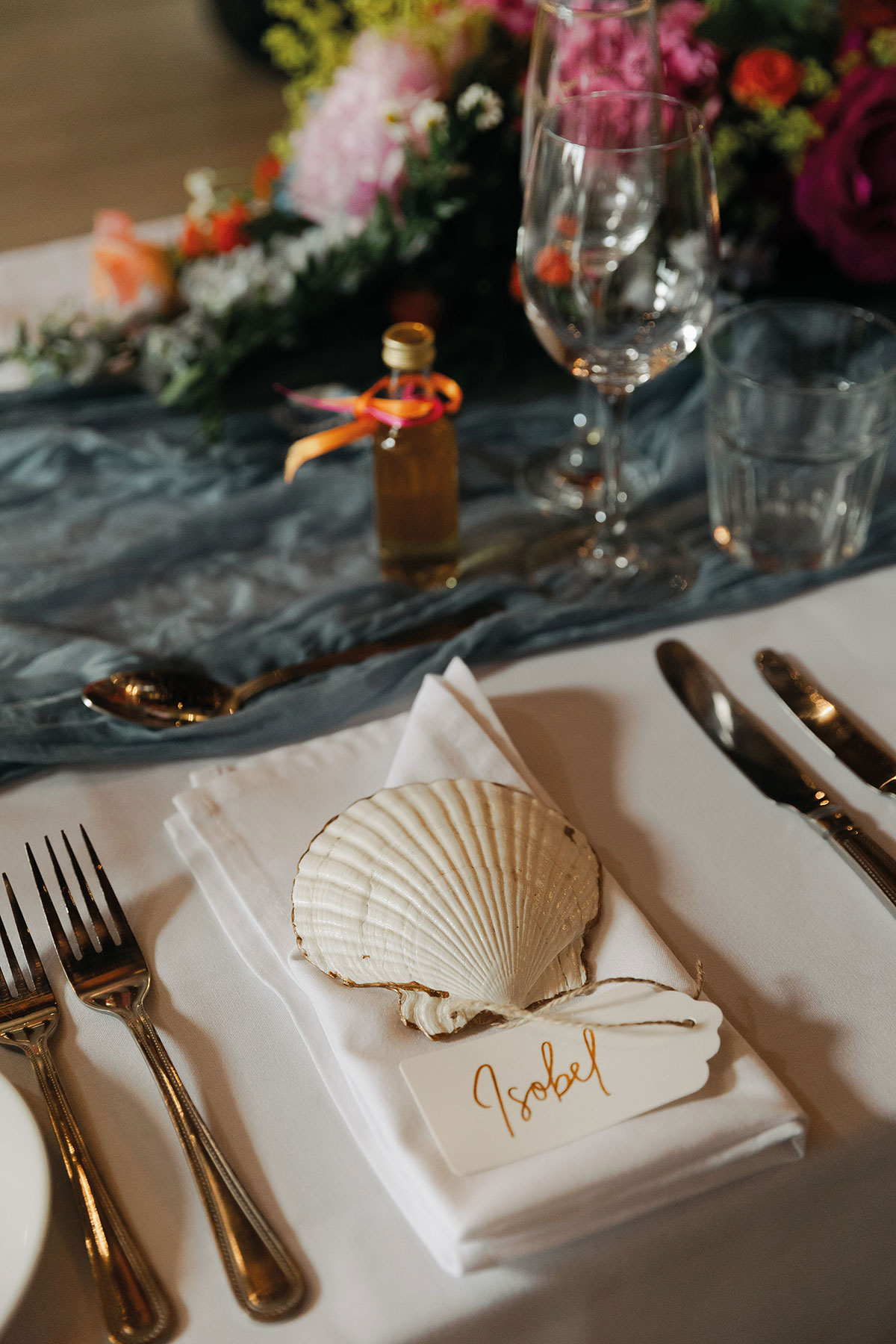 White shell place setting with handwritten name tag on a wedding tablescape with blue runner, flowers and glassware
