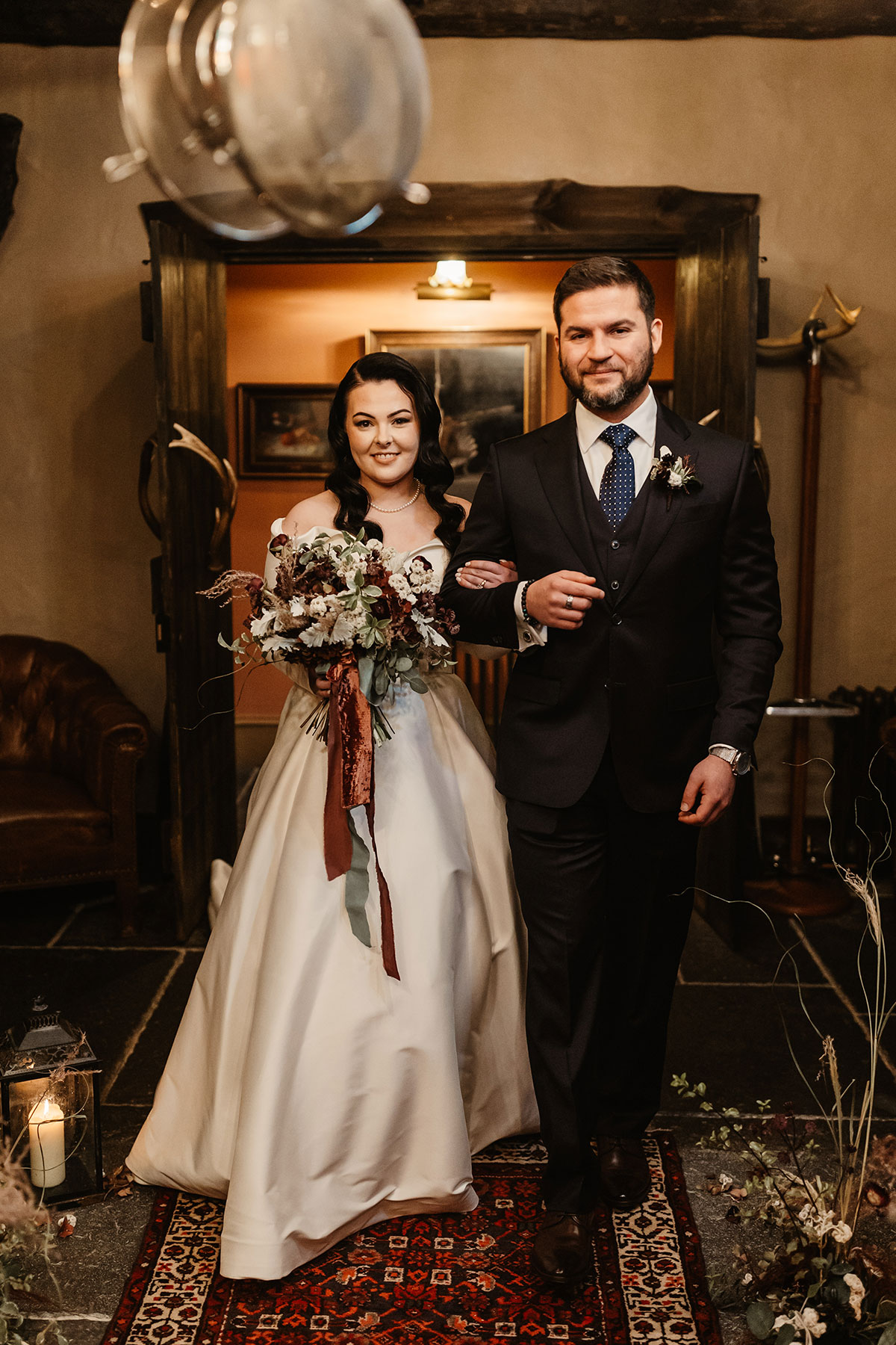 Bride and groom walking arm in arm down a patterned rug towards their ceremony space, surrounded by warm candlelight