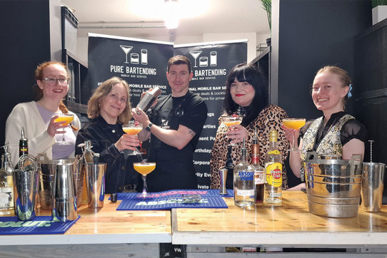 Four women holding glasses with cocktails surrounding one man holding cocktail shaker