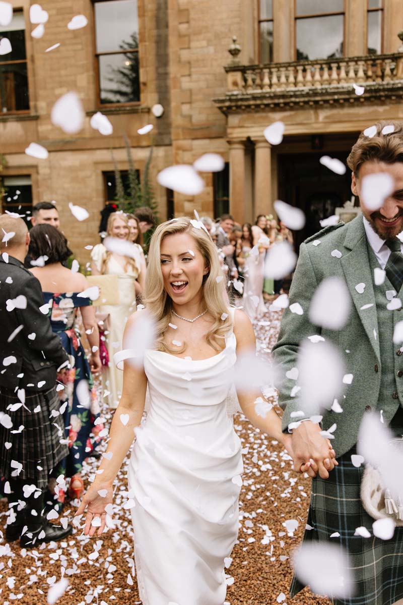Lia and Louis walk through white confetti outside Cambo House following their Fife wedding ceremony.