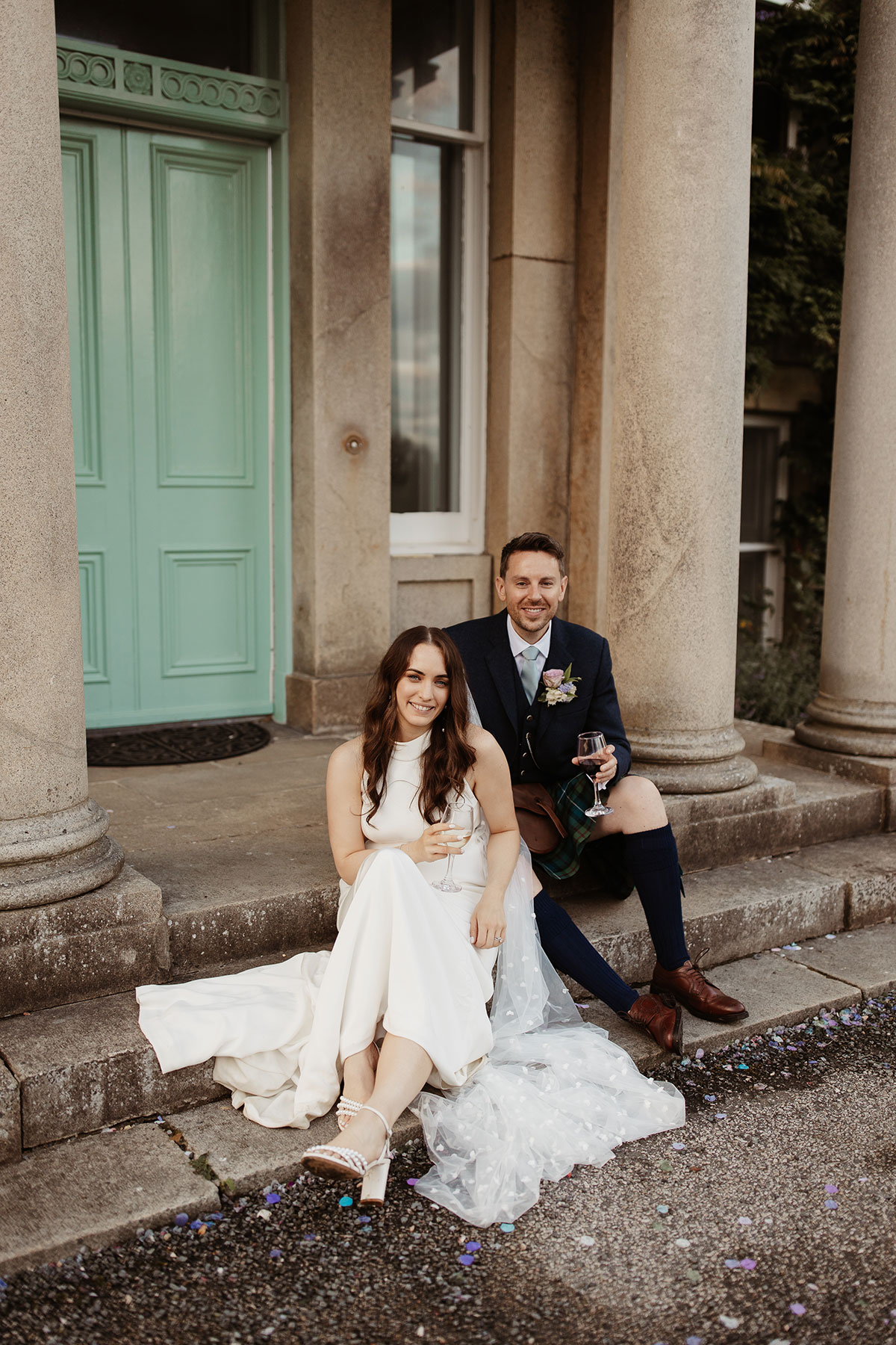 a bride and groom sitting on a set of stone steps in front of a turquoise door at their Netherdale House wedding