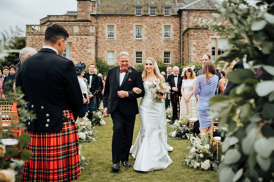 A person and person walking down an aisle in a garden with Archerfield House in the background.