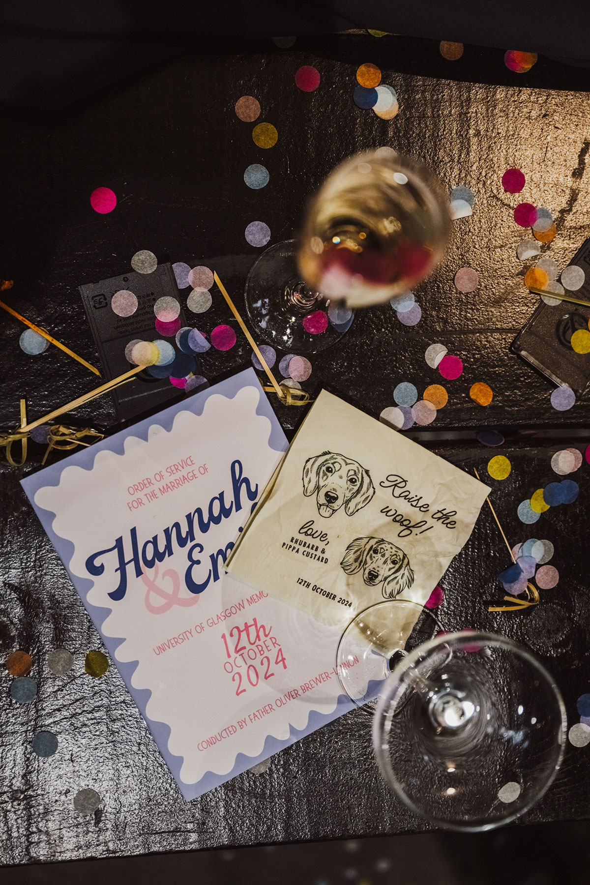 Flatlay of Hannah and Emily’s wedding order of service and personalised dog-illustrated napkin surrounded by scattered confetti and champagne glasses.