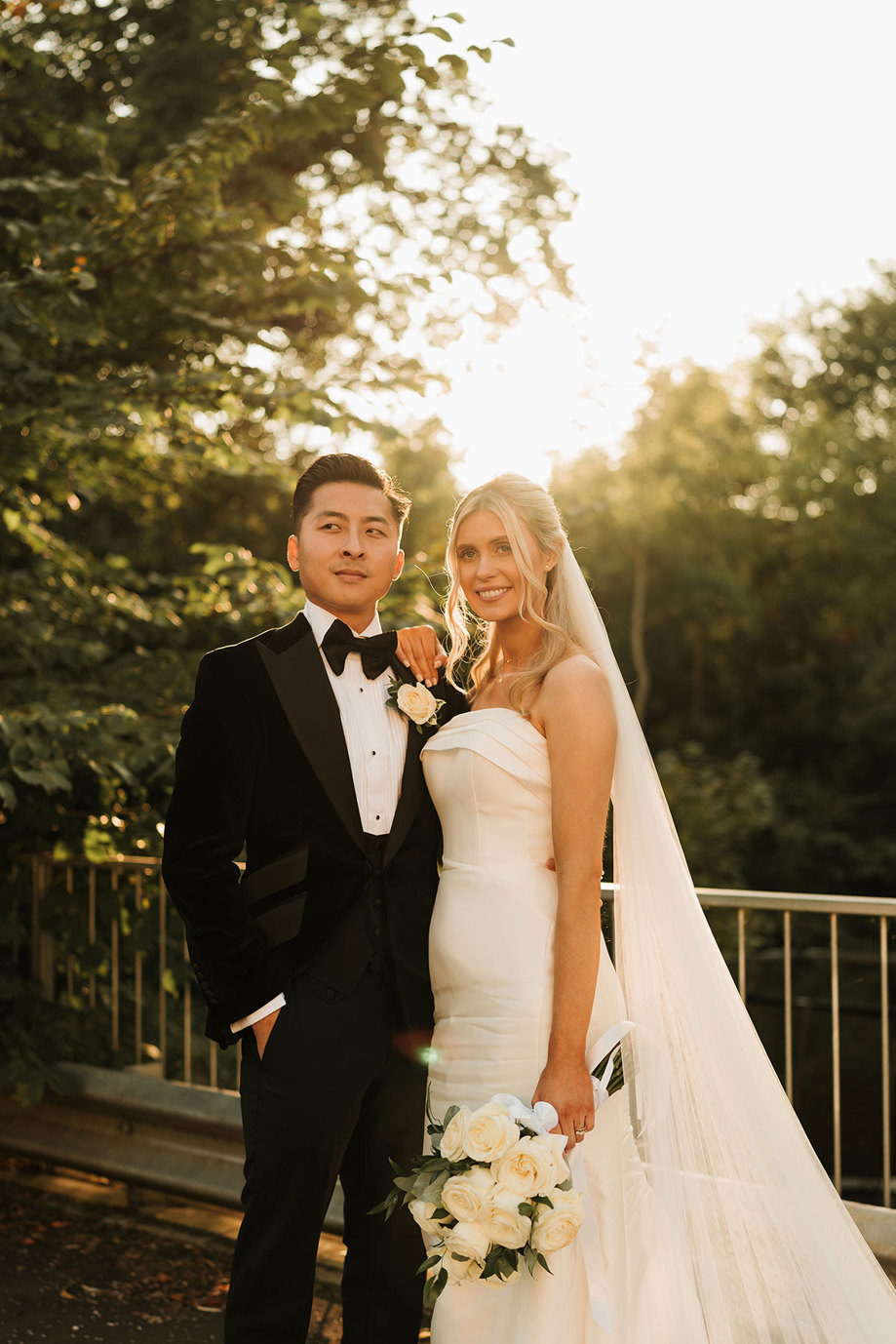 a bride and groom posing in soft sunlight in front of a metal rail and trees