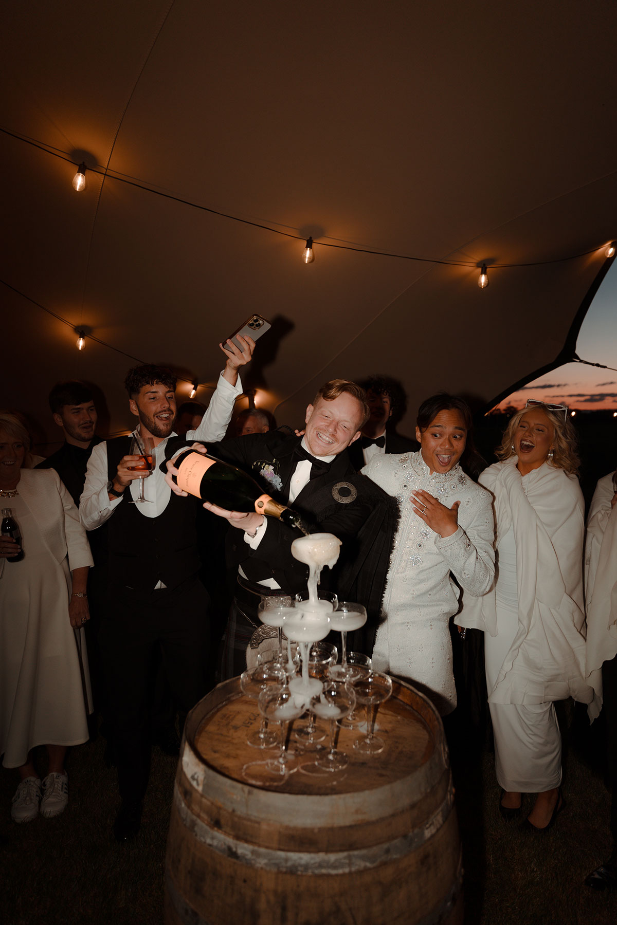 Grooms pouring champagne tower during evening reception under stretch tent at Falkirk farm wedding