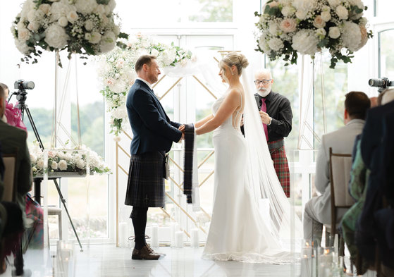 bride and groom hold hands during wedding ceremony with floral arrangements on either side