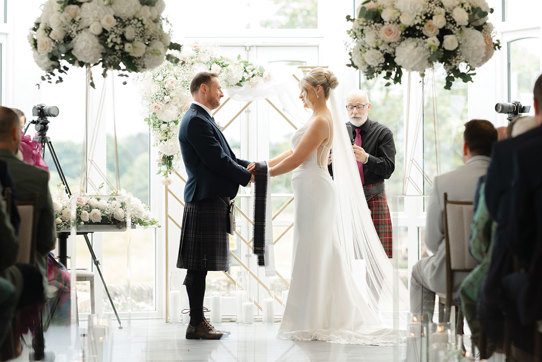 bride and groom hold hands during wedding ceremony with floral arrangements on either side