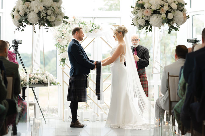 bride and groom hold hands during wedding ceremony with floral arrangements on either side