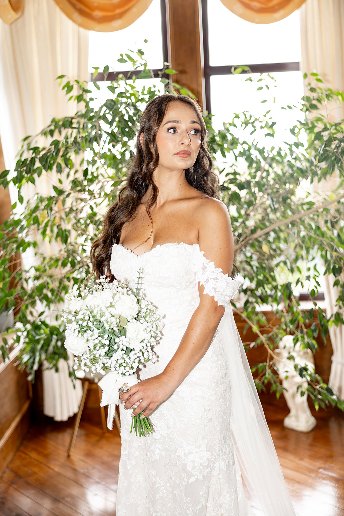 Bride in lace off-the-shoulder gown holds white bouquet by a window before her Falside Mill wedding ceremony