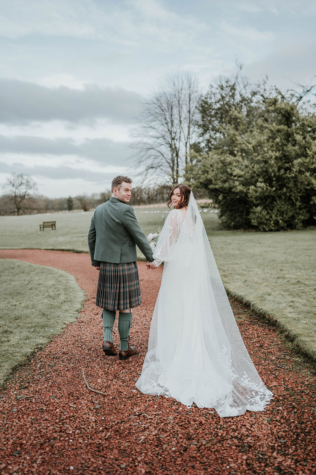Bride and groom looking back over their shoulders while walking hand in hand along a garden path