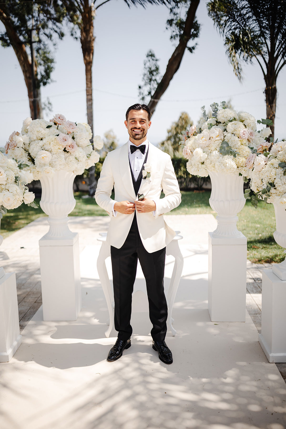 Groom in white tuxedo jacket and black bow tie stands between tall white floral arrangements outdoors