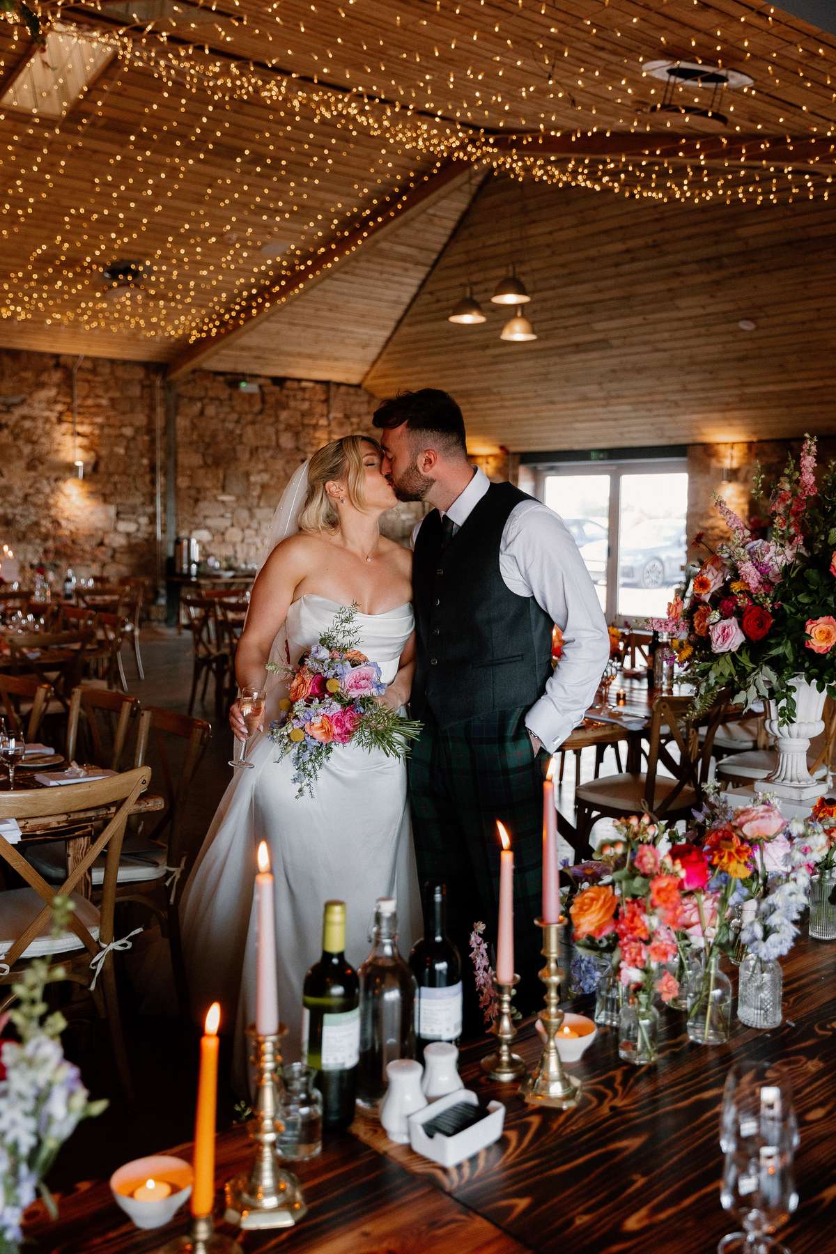 Bride and groom sharing a kiss in the candlelit reception space at Falside Mill with colourful floral styling