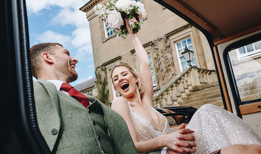 An elated bride sitting in a vintage wedding car outside Dumfries House wearing an ivory sequin wedding dress raising a bouquet aloft holding hands with a groom wearing a green tweed outfit