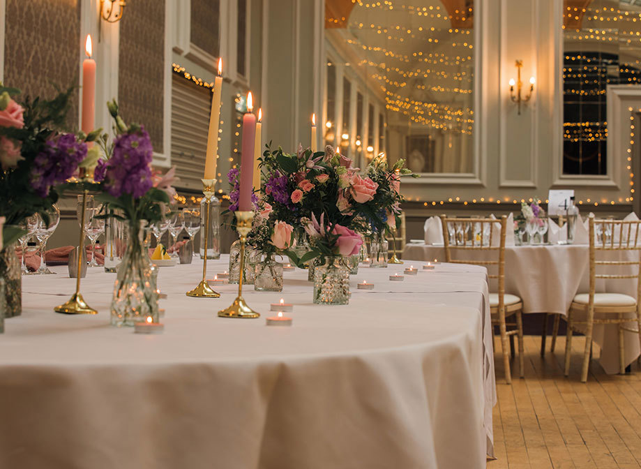 A round table in the foreground with a white table cloth and colourful candles and flowers, in the background are more tables and chair and lots of fairylights