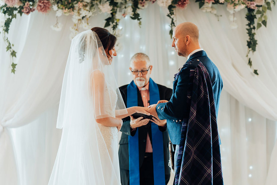 a groom wearing a tartan plaid places a ring on a bride wearing a veil. A minister stands between them looking downward at a book. There are soft white curtains and hanging floral decorations in background