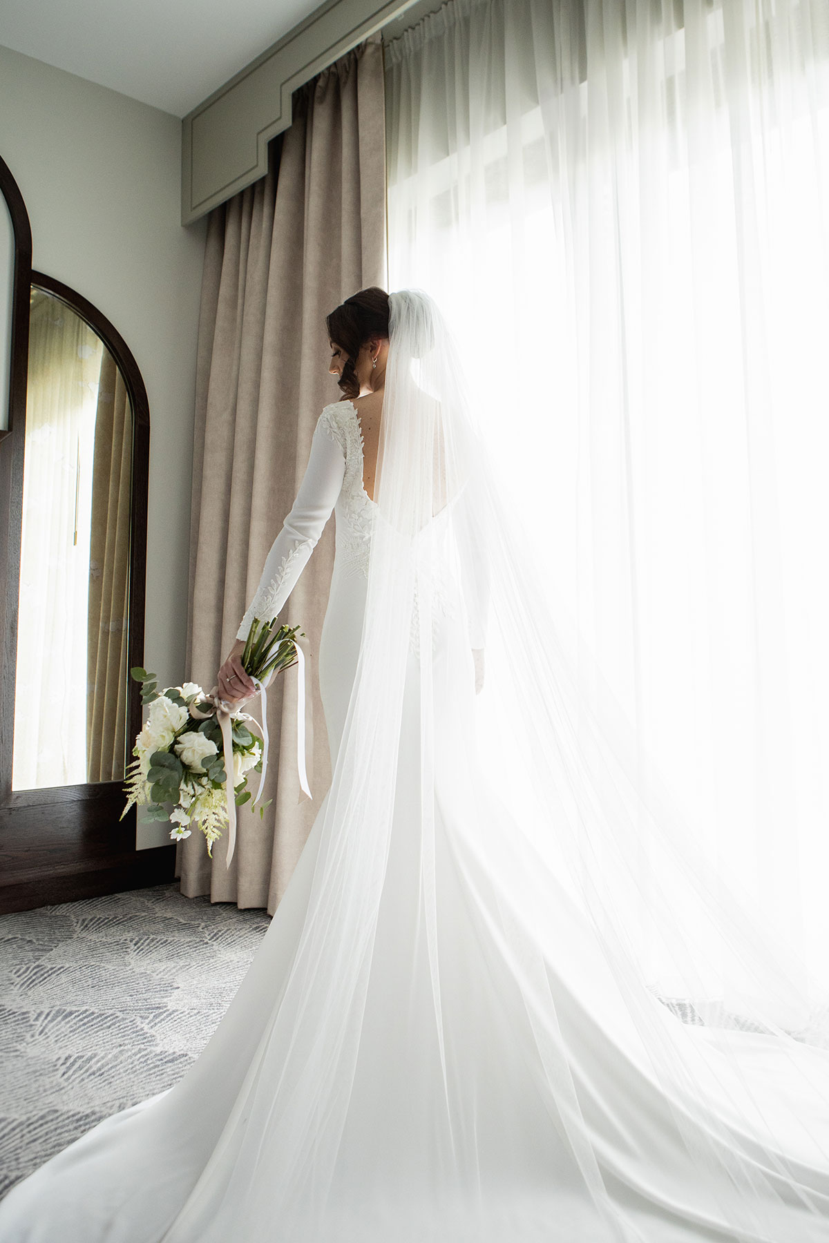 Bride in a fitted long-sleeved gown holding a bouquet, standing by a bright window with her veil flowing