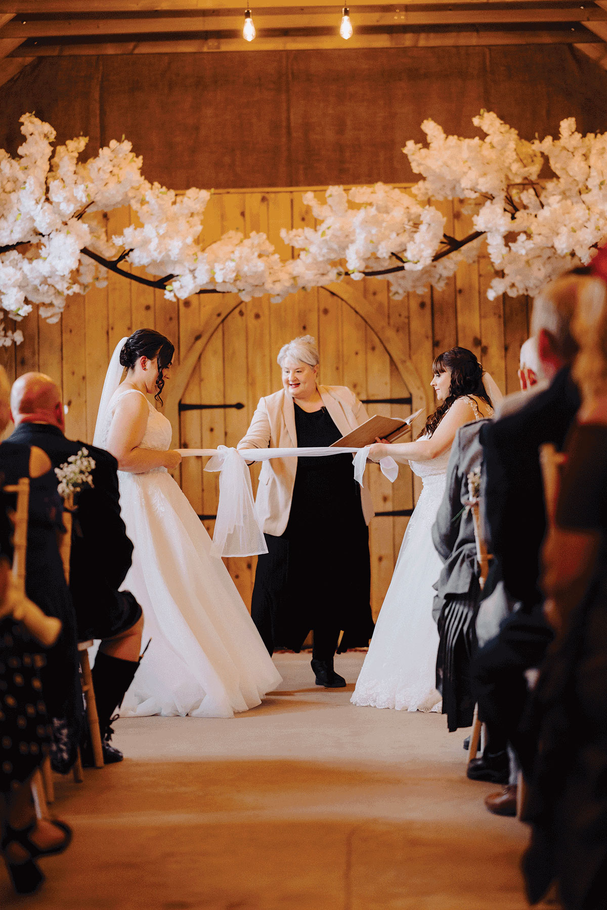 Two brides tying the knot at Den of Culross in a joyful same-sex wedding ceremony led by Fuze Ceremonies celebrant Yvonne beneath white blossom trees