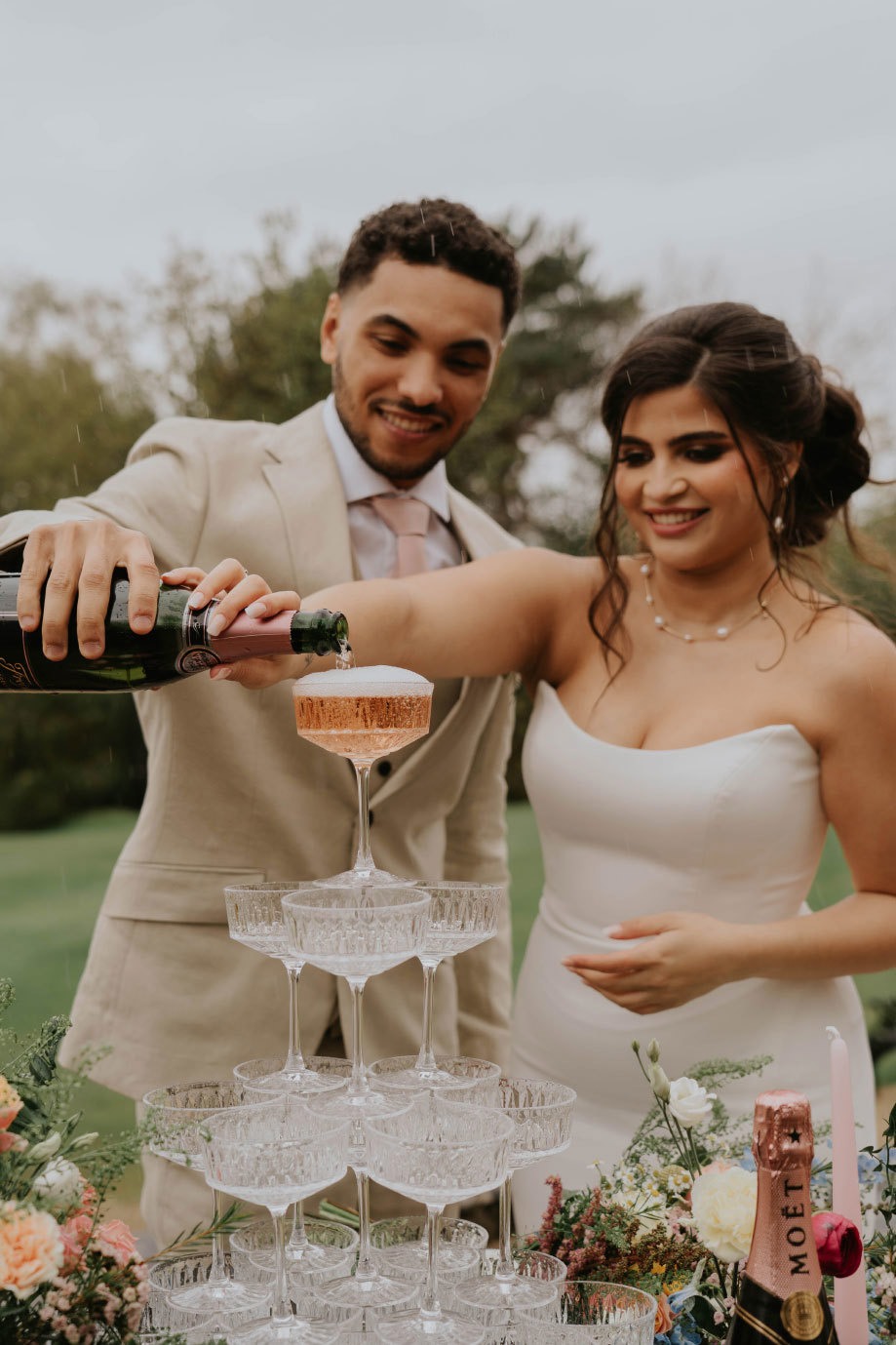 A bride and groom pour rose champagne into a champagne tower 