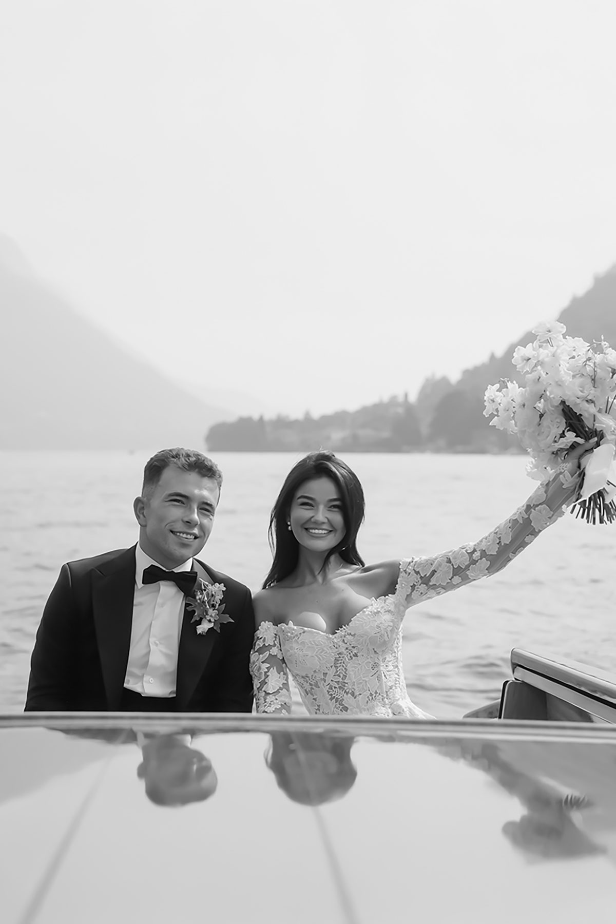 Bride and groom seated together on a classic Lake Como boat, with the lake and hillside villages behind them.