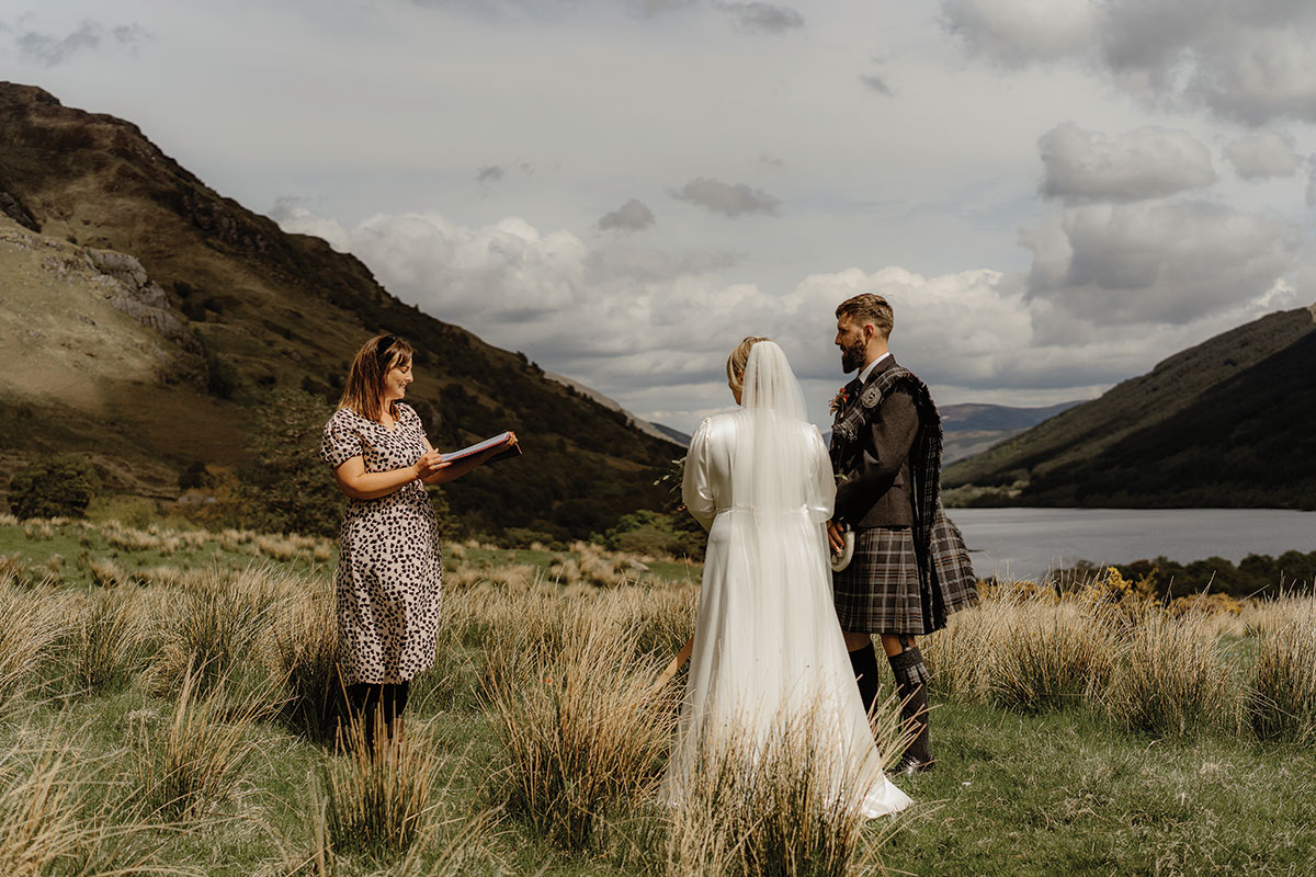 bride and groom hold hands overlooking the glistening Loch Doine near Balquhidder as celebrant conducts ceremony
