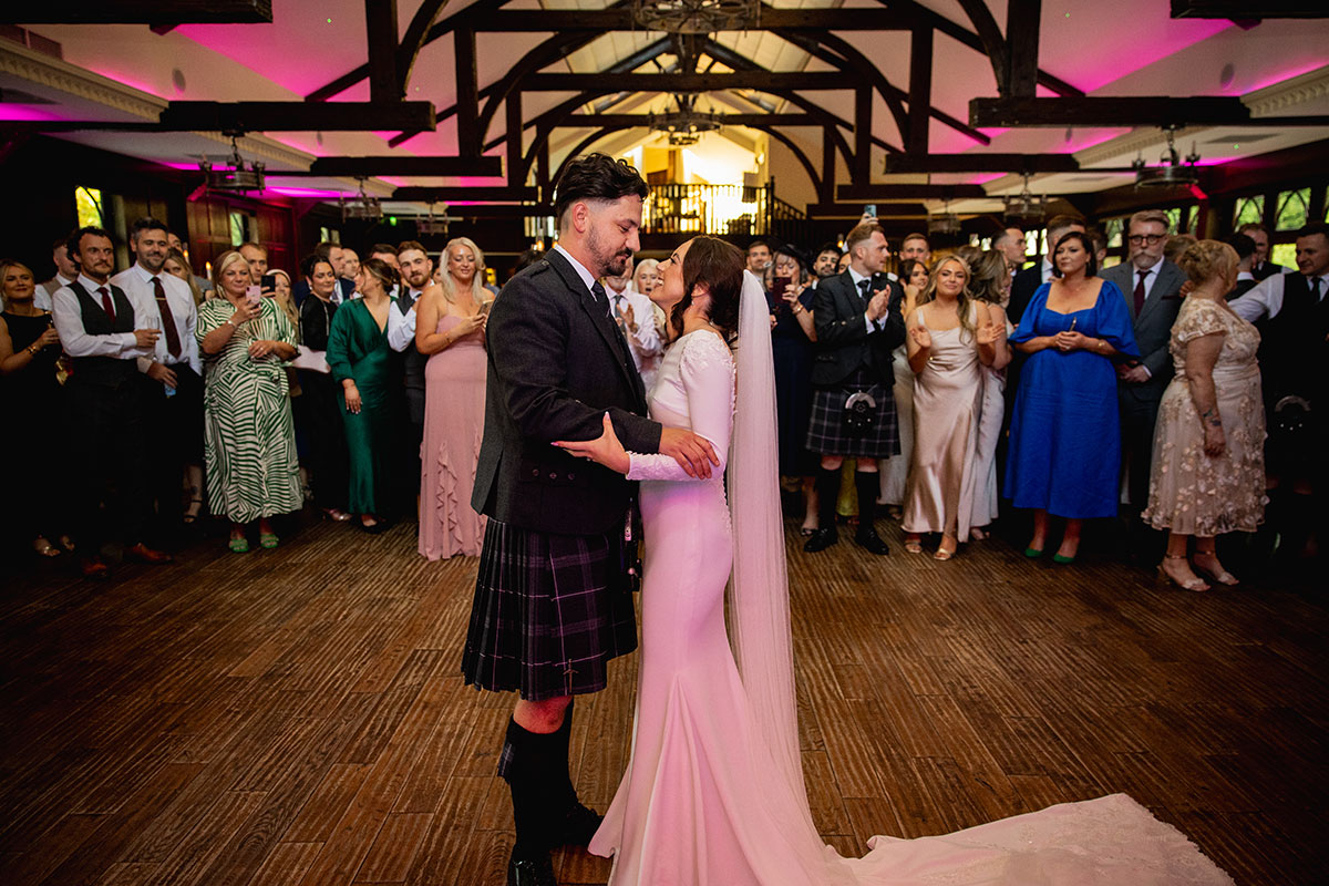Bride and groom share their first dance surrounded by cheering guests in an atmospheric hall with timber beams and pink lighting