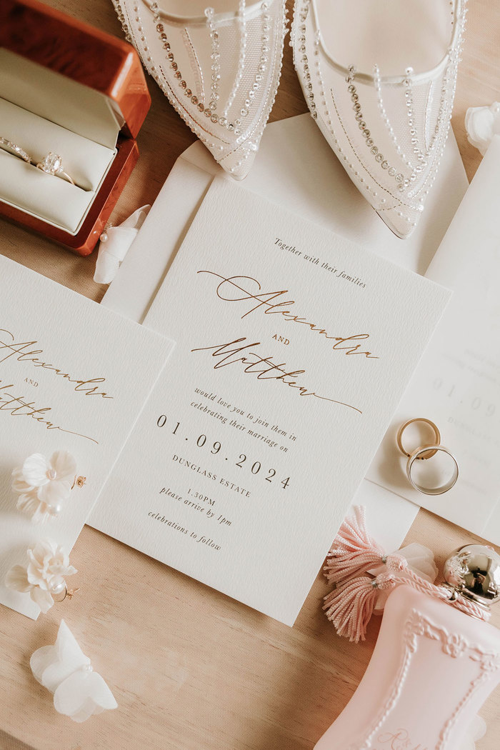 a wedding details flat lay of wedding stationery, shoes, rings, flower earrings and perfume bottle