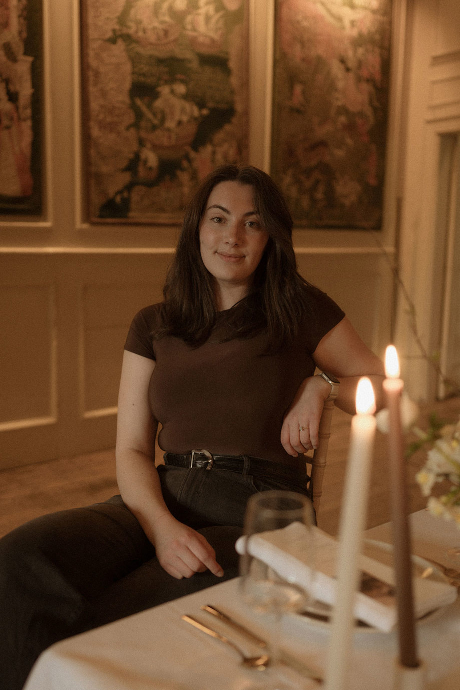dark haired woman in brown t-shirt and black trousers smiles softly, sitting in a chair alongside the a set dining table, complete with a white tablecloth and lit candlesticks